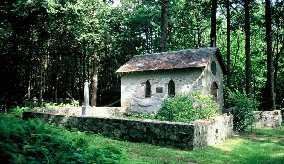 A small stone chapel surrounded by a short stone wall, green grass and bushes and a lush green forest in the background.