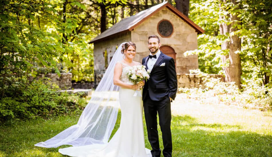 A wedding couple in front of a stone chapel surrounded by lush green trees and woods.
