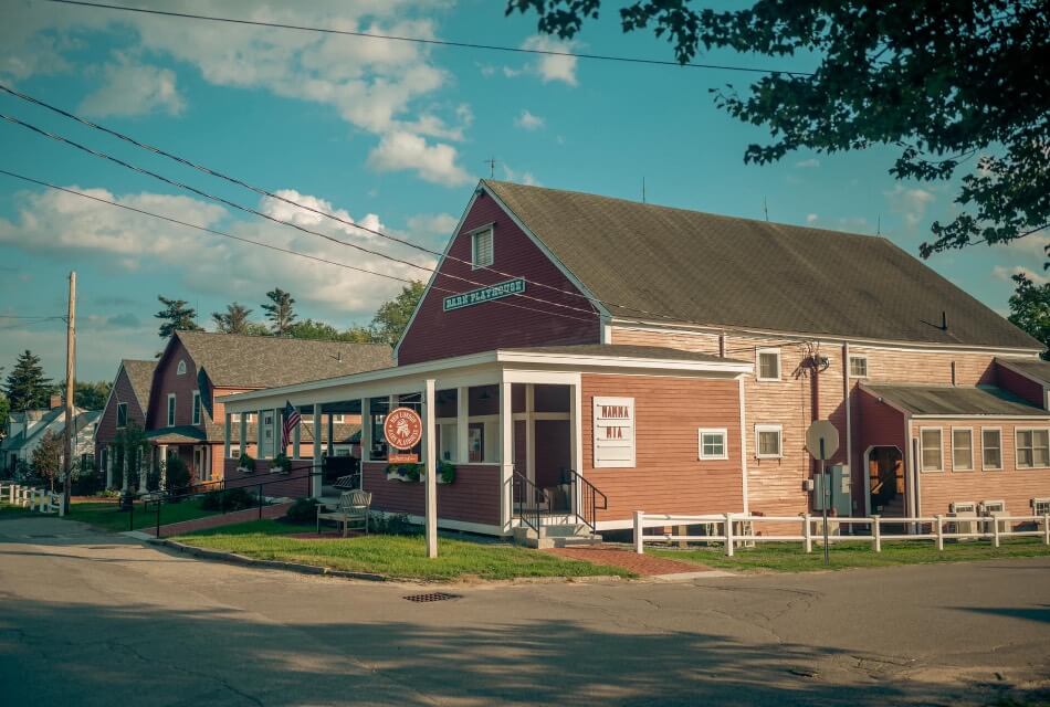 A brick building with white trim and a covered porch, with green grass in front and mountains in the background.