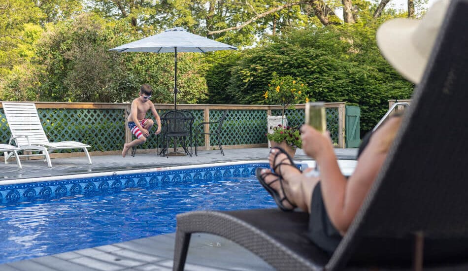 a woman lounging by a pool watching a boy jumping into the pool.
