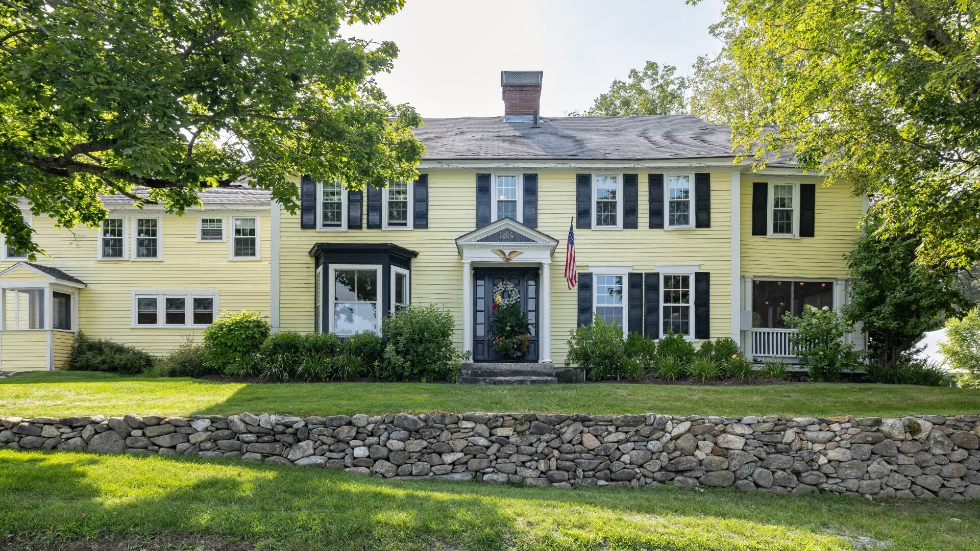 A stately 2-story yellow house with white trim with green grass and surrounding trees.