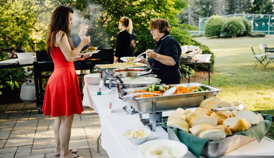 Catering outside with long tables with white tablecloths, chafer dishes with food, people serving and a woman in line getting food.