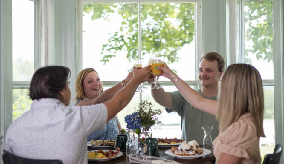 Four people sitting at a breakfast table lifting glasses of orange juice and toasting.