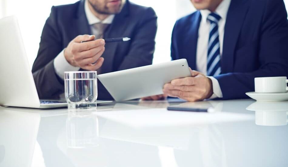 2 men in blue suits discussing items over a tablet and laptop with a glass of water and a cup of coffee.