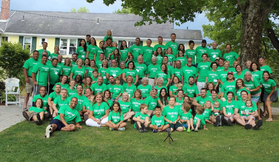 A family reunion with lots of people wearing matching green t-shirts sitting and standing on the grass in front of a large yellow and white house.