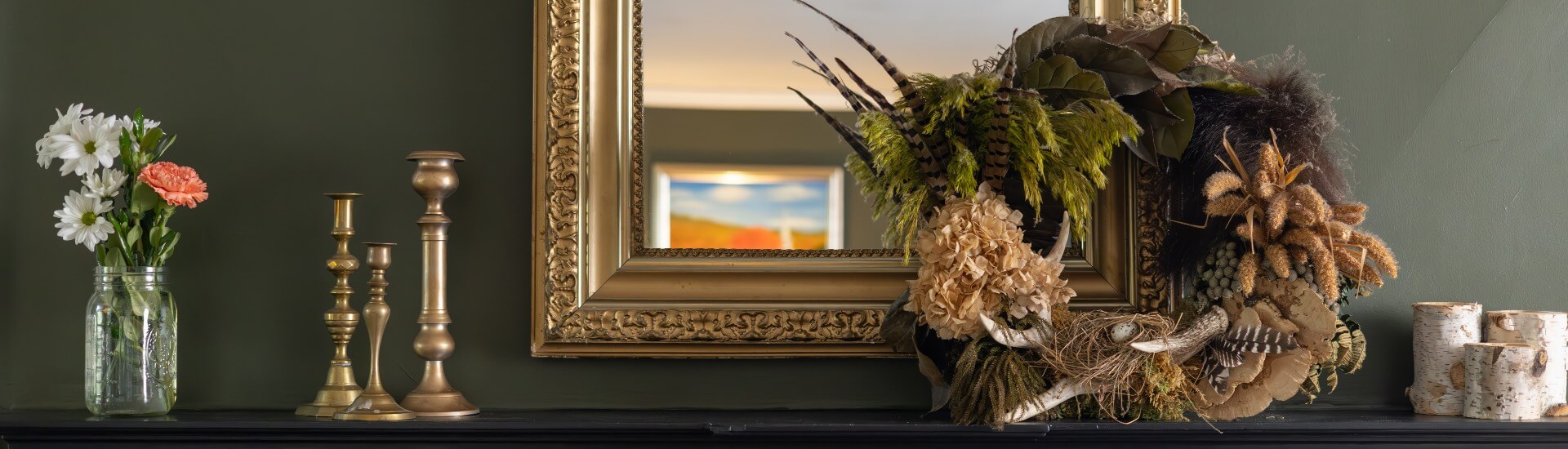 A mantle with a wreath, candlesticks, flowers, and blocks of birch trees on the mantle, and an ornate mirror above the mantle.