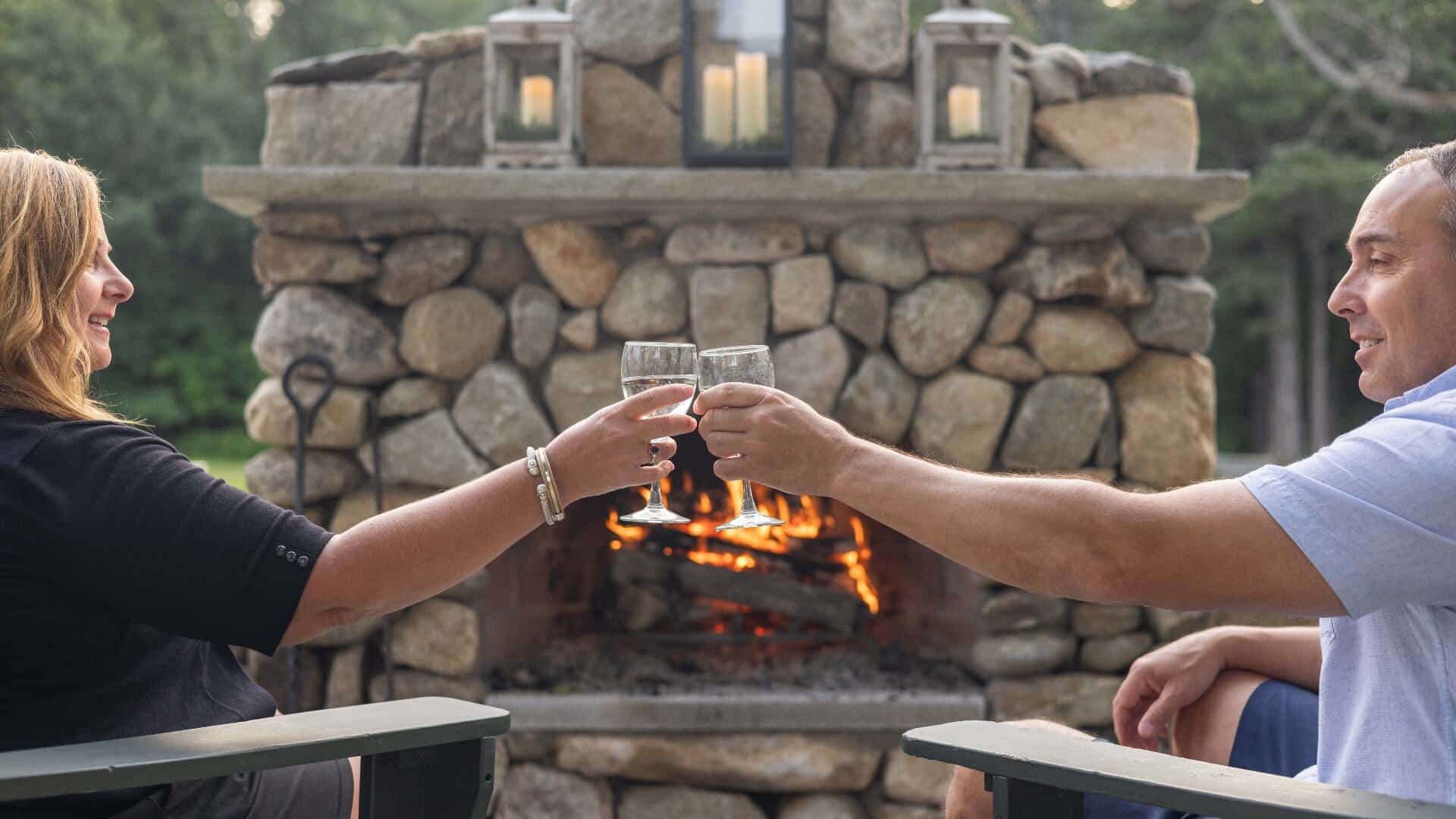 A couple toasting with glasses of white wine, sitting in wood armchairs outside by a stone fireplace with a fire going.