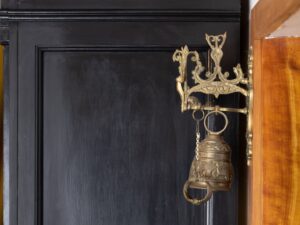 An ornate brass bell and wall holder mounted on a wood wall on the side of a black cupboard.
