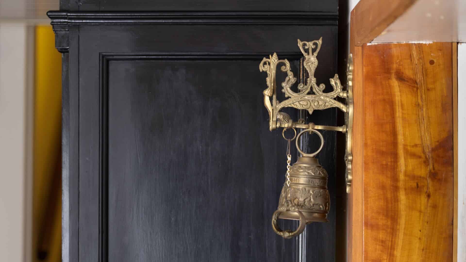 An ornate brass bell and wall holder mounted on a wood wall on the side of a black cupboard.