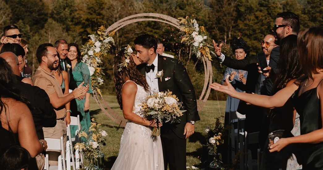 A wedding couple kissing after saying their vows amidst onlookers, in front of sweeping mountains and trees.