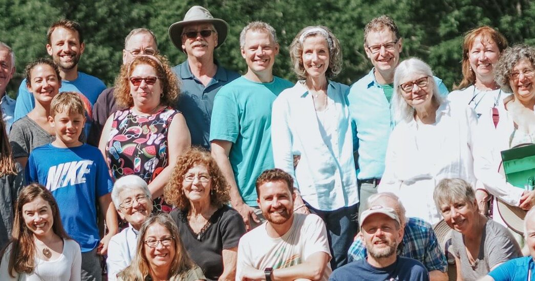 A group of smiling people at a family reunion, ranging from grandparents down to babies, in front of green trees and bushes.