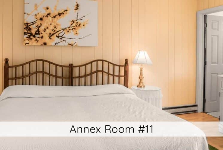 A bedroom with yellow walls, a bed with a wood headboard, white bedding, round tables with lamps on either side of the bed, and a sink with mirror above it along one wall. Titled Annex Room #11.
