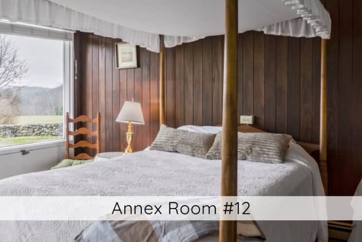 A bedroom with a canopy bed with wood wall behind the bed, small tables with lamps on either side of the bed, a wood chair in the corner, and a large picture window with beautiful views of trees and the mountains. Titled Annex Room #12.