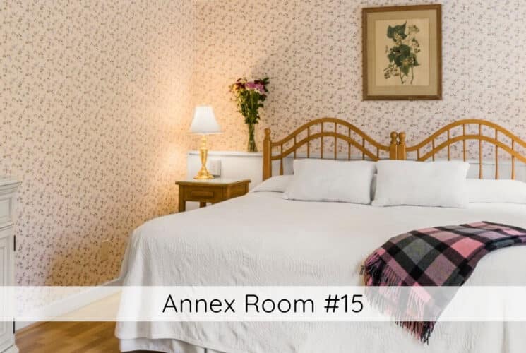 A bedroom with floral wallpaper, wood floors, a bed with a wood headboard and white bedding with a plaid throw, nightstands with lamps on either side of the bed, and a sink with a mirror above it on one wall. Titled Annex Room #15.
