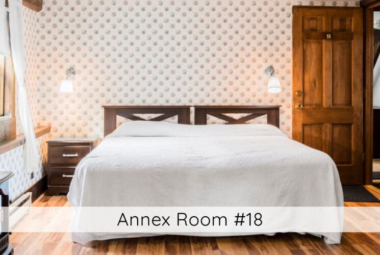 A bedroom with floral wallpaper, a bed with wood headboard, white bedding, a nightstand on one side of the bed, a dresser and electric fireplace, wood floors, a window on 1 of the walls with white curtains. Titled Annex Room #18.