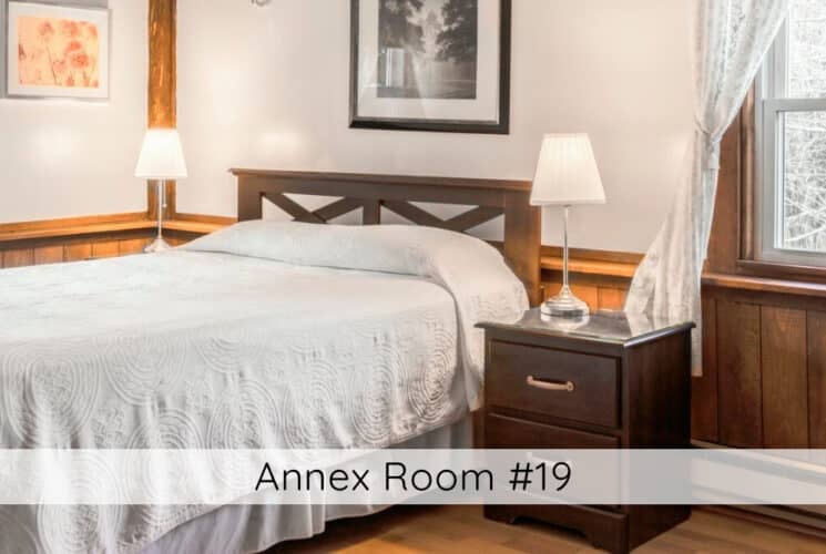 A bedroom with a bed with wood headboard, white bedding, nightstands with lamps on either side of the bed, an armchair in one corner, wood floors and wainscotting, and windows on 2 of the walls with white curtains. Titled Annex Room #19.