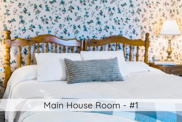 A bedroom with floral wallpaper, a bed with wooden headboard, white bedding, an grey accent pillow and plaid throw, nightstands with lamps on either side of the bed. Titled Main House Room #1.