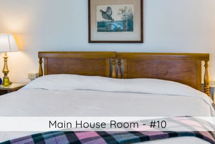 A bedroom with a bed with wood headboard, white bedding, colorful throw on the bed, nightstands on either side of the bed with lamps, and photos of birds on the wall. Titled Main House Room #10