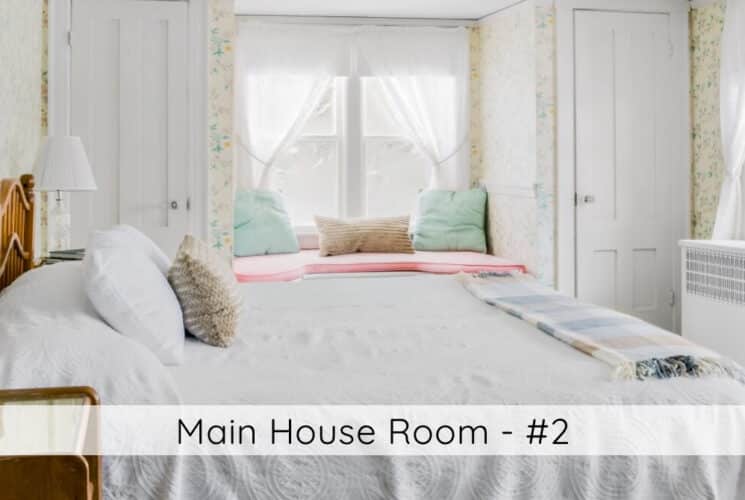A bedroom with a bed with wood headboard and white bedding, nightstands with lamps on either side of the bed, an antique dresser and mirror on the wall opposite the bed, and a sitting bench tucked into a large window alcove. Titled Main House Room #2