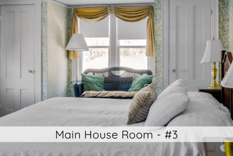 A bedroom with a bed with wood headboard, nightstands with lamps on either side of the bed, a window with a wicker loveseat in front of the window, and a heater on the other wall in front of the window with a vase of flowers on the heater. Titled Main House Room #3.