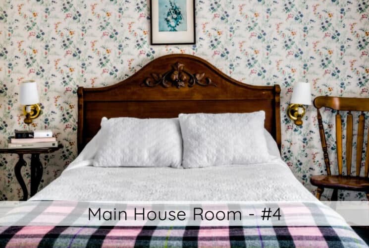A bedroom with colorful floral wallpaper, a bed with a high wood-carved headboard, white bedding with a plaid throw at the foot of the bed, a small table on one side of the bed with books, lamps on either side of the bed on the wall, a wood chair next to the other corner of the bed, and a window with white curtains. Titled Main House Room #4