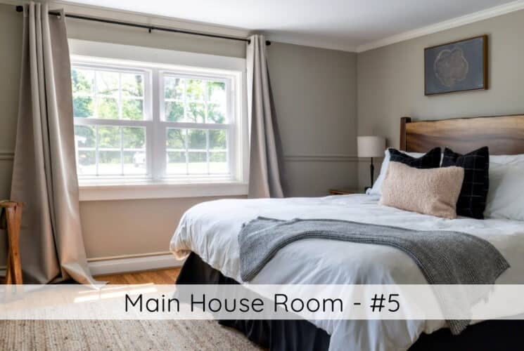 A bedroom with a rustic wood headboard, white bedding with grey throw and colored accent pillows, nightstands with lamps on either side of the bed, a white wardrobe in the corner, wood floors, grey and tan walls, satin curtains on either side of a large window. titled Main House Room #5.