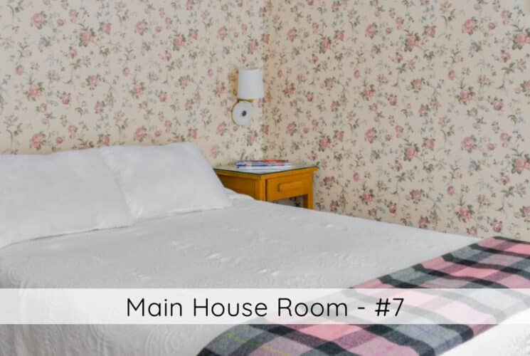 A bedroom with flowered wallpaper, a bed with a wood headboard, white bedding with a plaid throw at the foot of the bed, a nightstand in the corner, and 2 lamps hanging on either side of the bed. Titled Main House Room #7