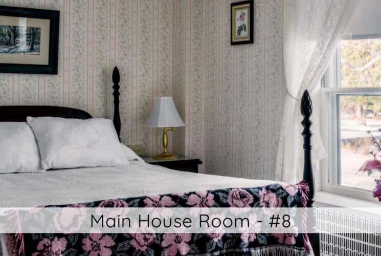 A bedroom with soft wallpapered walls, a fourposter bed, white bedding, a colorful flowered throw at the foot of the bed, an open window and flowers on the window sill, titled Main House Room #8.