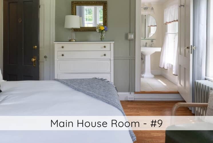 A bedroom with a bed, armchair, white dresser and mirror, a window, a door open to a bathroom, and titled Main House Room #9