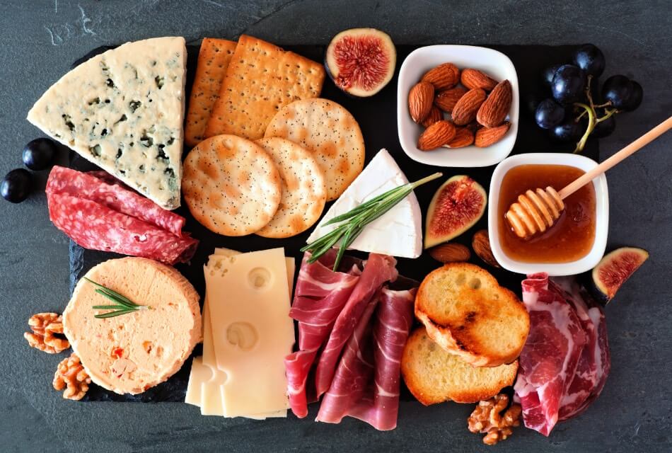 A black slate charcuterie tray with varieties of cheeses, meats, nuts, fruit, honey, crackers, and bread.