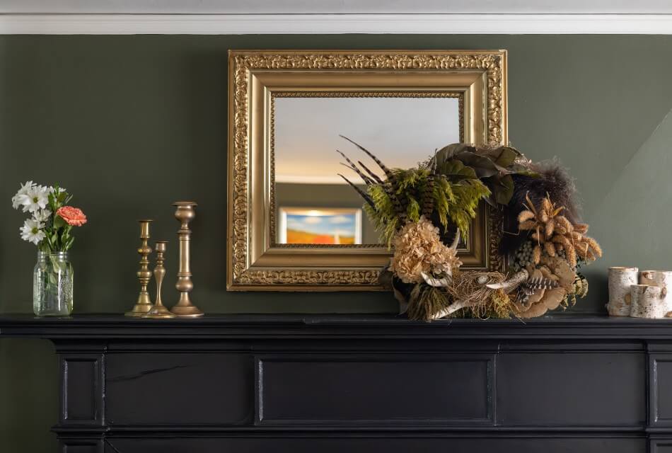 A mantle with a wreath, candlesticks, flowers, and blocks of birch trees on the mantle, and an ornate mirror above the mantle.