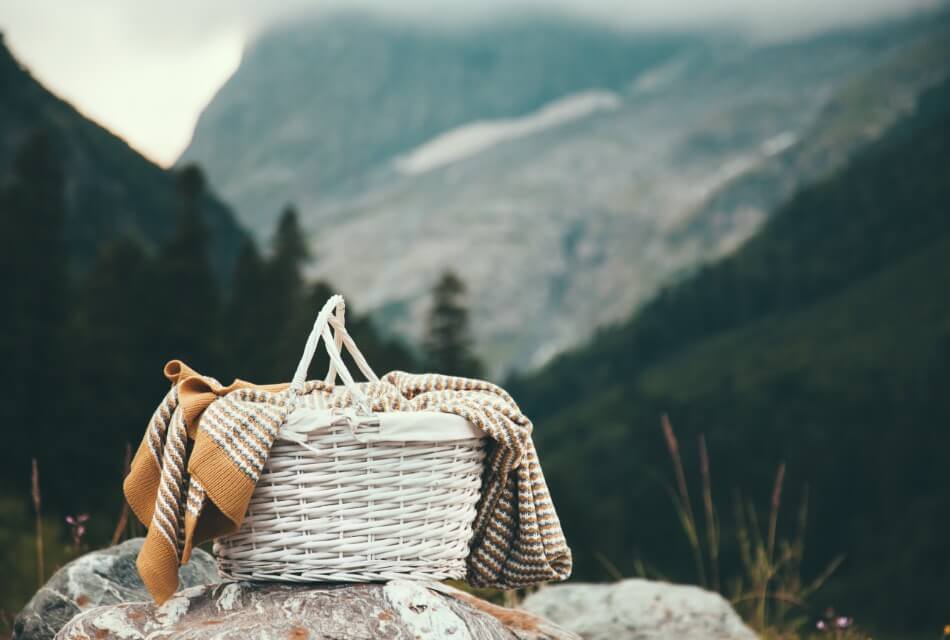 A white wicker picnic basket with a multi colored tea towel, set on rocks with mountains in the background.