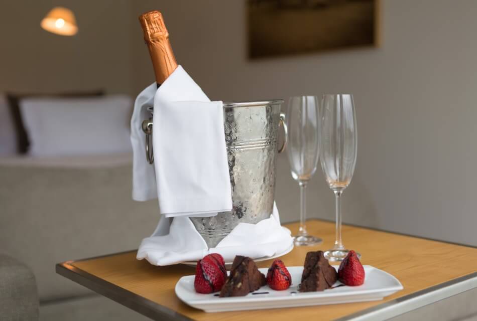 A bucket of champagne with a white cloth, two champagne glasses, and a white plate with strawberries and pieces of chocolate brownies drizzled with chocolate sauce.