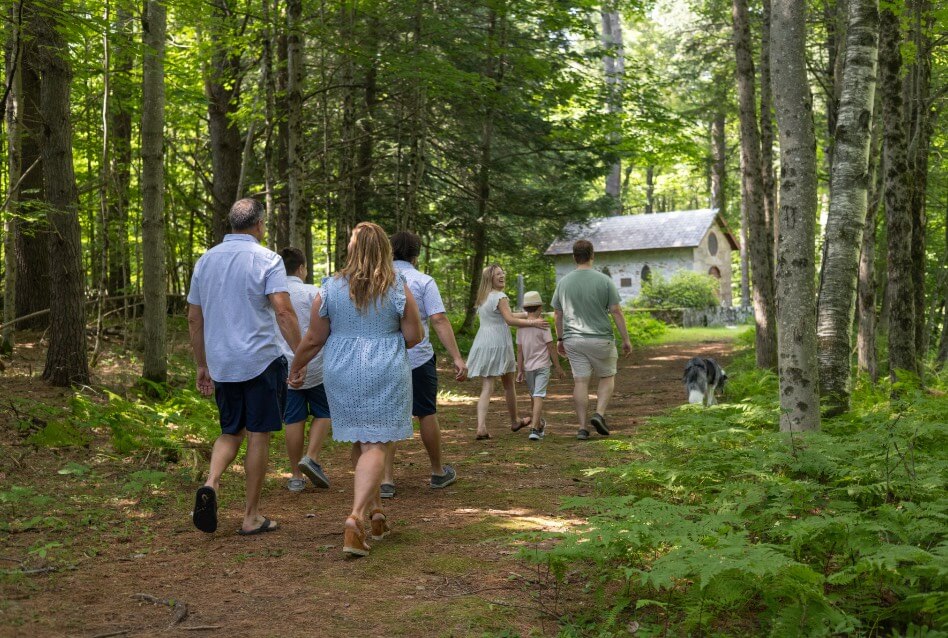 A group of people and a dog walking through the woods towards an outdoor stone chapel.