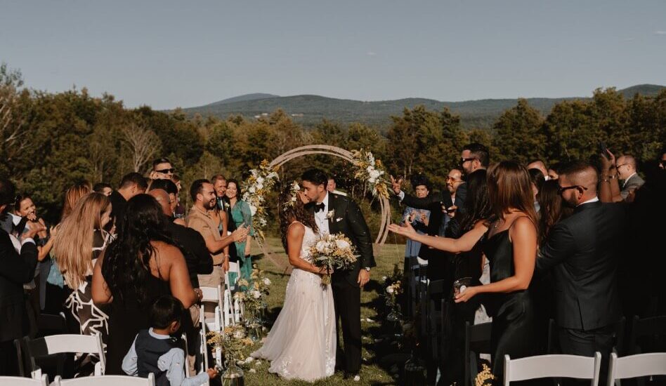 A wedding couple kissing after saying their vows amidst onlookers, in front of sweeping mountains and trees.