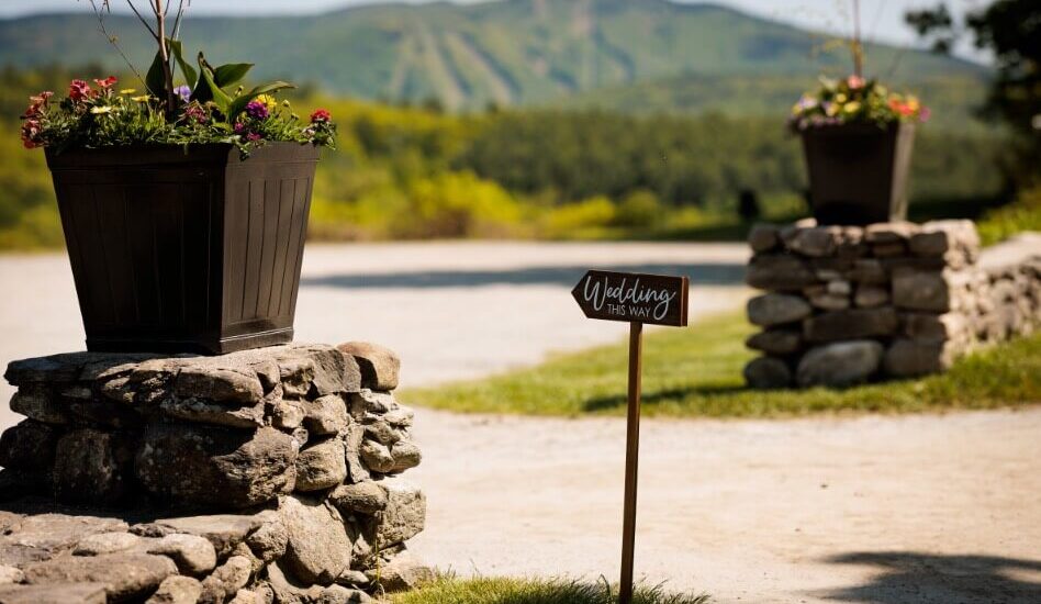 An outdoor area with two rock walls topped with flower boxes, and sweeping mountains in the background, and a sign near one of the walls that says Wedding This Way.